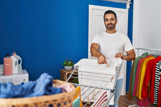 Nuori latino mies hymyilee luottavainen roikkuu vaatteita pyykkinarulla klo pesutupa - Valokuva, kuva