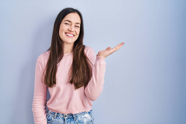 Young brunette woman standing over blue background smiling cheerful presenting and pointing with palm of hand looking at the camera.  - Photo, Image