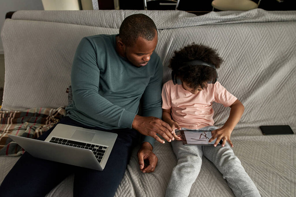 Top view of father with laptop look at little son draw on digital tablet on sofa at home. Young black family lifestyle and relationship. Domestic leisure and entertainment. Fatherhood and parenting - Foto, Imagem