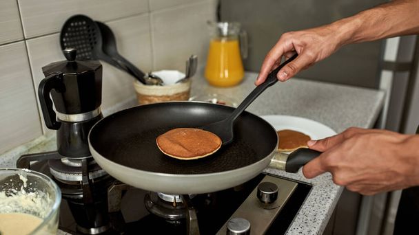 Cropped view of man taking pancake with kitchen spatula after cooking on pan on electric stove at home kitchen. Concept of domestic lifestyle. Idea of healthy eating. Modern apartment. Daytime - Foto, afbeelding