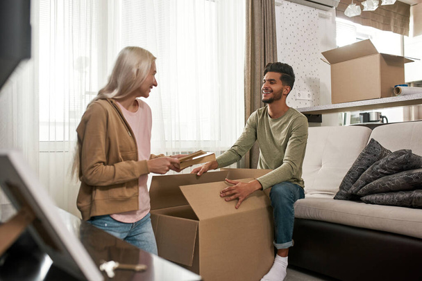 European girl packing books in cardboard box and looking at her middle eastern husband or boyfriend at living room. Young smiling multiracial couple moving to new apartment. Concept of home relocation - Фото, зображення