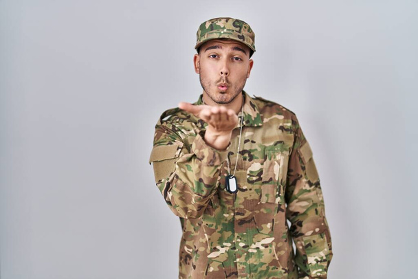 Jovem hispânico vestindo uniforme do exército de camuflagem olhando para a câmera soprando um beijo com a mão no ar sendo adorável e sexy. expressão do amor.  - Foto, Imagem