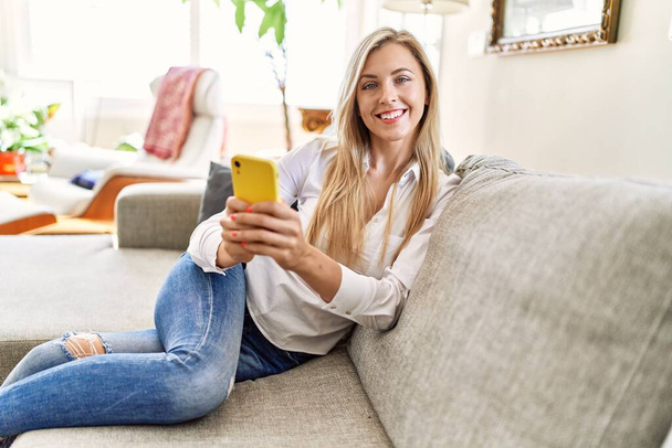 Νεαρή ξανθιά γυναίκα που χρησιμοποιεί το smartphone κάθεται στον καναπέ στο σπίτι. - Φωτογραφία, εικόνα