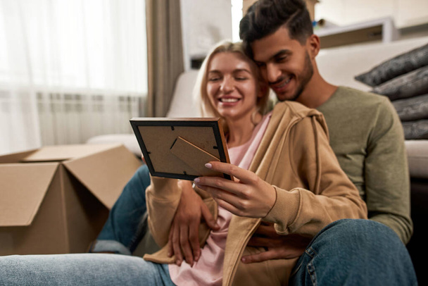 Focus on foreground of photo looking blurred young smiling multiracial couple on floor during packing things in cardboard box. Man and woman moving to new apartment. Home relocation. Family memories - Φωτογραφία, εικόνα