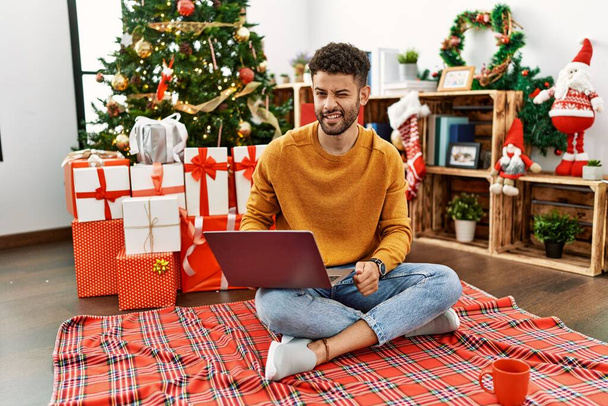 Arab young man using laptop sitting by christmas tree winking looking at the camera with sexy expression, cheerful and happy face.  - Fotó, kép