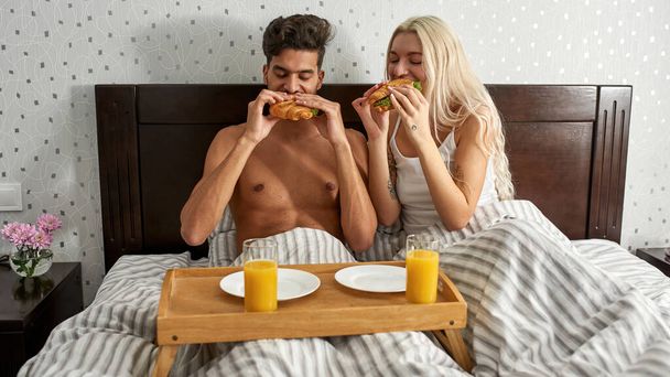 Young multiethnic couple eating sandwiches during breakfast on bed at home. Caucasian girl and middle eastern man. Domestic lifestyle. Rest at home. Spacious bedroom in modern apartment. Morning time - Foto, Bild