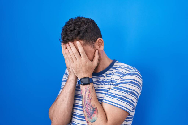 Νεαρός Ισπανός στέκεται πάνω από το μπλε φόντο με λυπητερή έκφραση που καλύπτει το πρόσωπο με τα χέρια ενώ κλαίει. έννοια της κατάθλιψης.  - Φωτογραφία, εικόνα