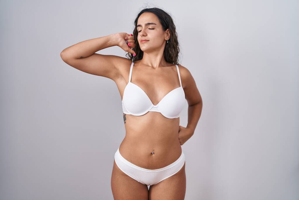 jonge Spaanse vrouw dragen witte lingerie stretching terug, moe en ontspannen, slaperig en gapen voor de vroege ochtend  - Foto, afbeelding