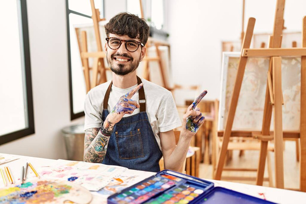 Homme hispanique avec barbe au studio d'art souriant et regardant la caméra pointant avec deux mains et doigts sur le côté.  - Photo, image
