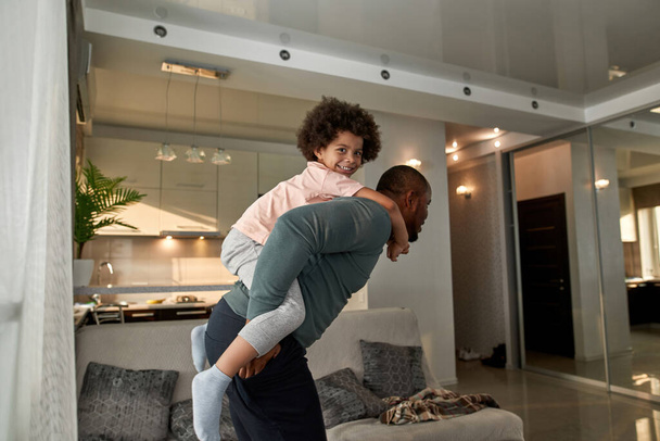 Side view of father carrying his little son piggyback at home. Smiling black man and child playing and enjoying time together. Fatherhood and parenting. Domestic leisure and entertainment - Foto, Imagen