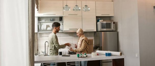 Side view of caucasian girl cutting air bubble film with scissors for packing bowl in hands of middle eastern man at kitchen. Young multiethnic couple moving to new apartment. Home relocation - 写真・画像