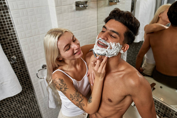 Kaukasische Mädchen rasieren den Bart ihres nahöstlichen Freundes morgens im Badezimmer mit Rasiermesser. Häuslicher Lebensstil. Gesichtspflege. Junge multiethnische Paarbeziehung. Mann und Frau lächeln - Foto, Bild