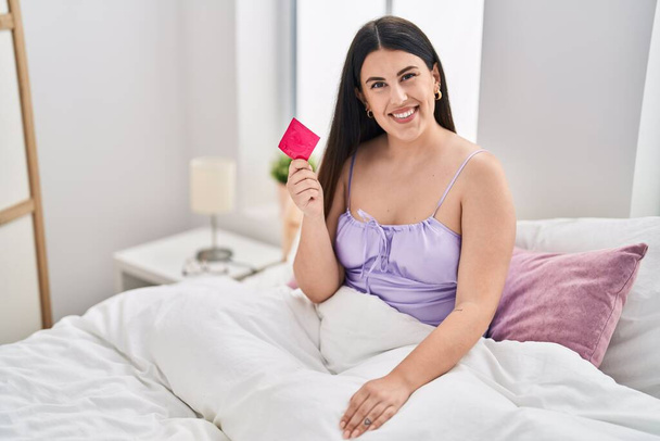 Giovane donna bruna che indossa lingerie e tiene il preservativo sul letto guardando positivo e felice in piedi e sorridendo con un sorriso fiducioso mostrando i denti  - Foto, immagini
