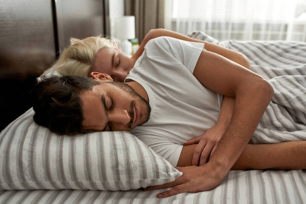 Side view of young multiethnic couple sleeping under blanket on bed. Caucasian girl hugging middle eastern boyfriend. Domestic lifestyle. Rest at home. Spacious bedroom in modern flat. Morning time - 写真・画像