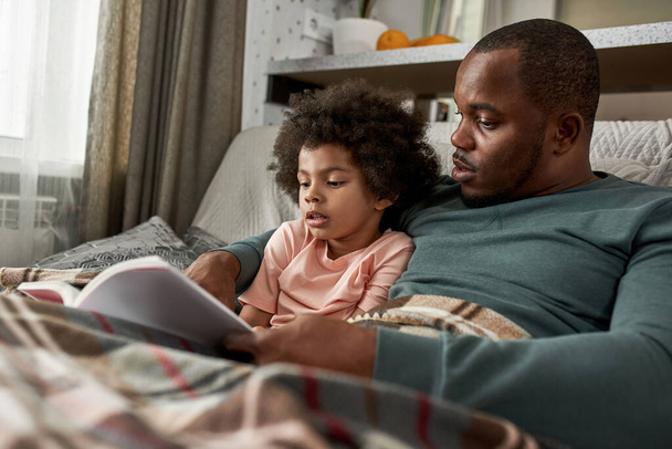 Focused adult father and little son reading book on sofa under blanket at home. Young black family spending time together. Fatherhood and parenting. Domestic leisure, hobby and entertainment - Foto, Imagem