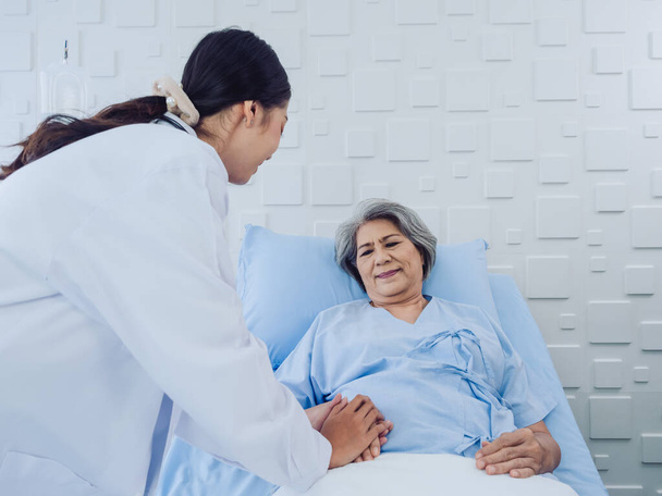 Счастливая пожилая азиатская пациентка в светло-голубом платье, лежащем на кровати в солевом растворе, была воодушевлена молодой женщиной-врачом в белом костюме со стетоскопом и держась за руку в больничной палате. - Фото, изображение