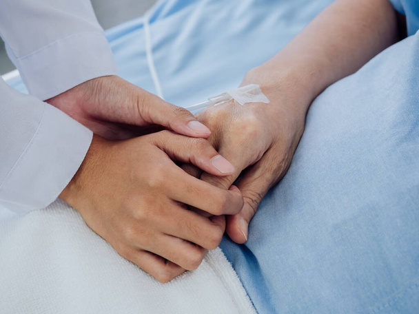 Κοντινό πλάνο γυναίκα γιατρός σε λευκό κοστούμι κρατά το χέρι της ηλικιωμένης ηλικιωμένης γυναίκας ασθενή σε γαλάζιο φόρεμα με ένα αλατούχο σωλήνα που βρίσκεται στο κρεβάτι του νοσοκομείου, ενθαρρύνοντας την έννοια του ασθενούς. - Φωτογραφία, εικόνα