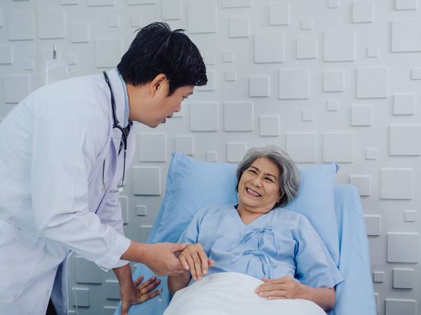 Le gentil médecin asiatique en costume blanc rend visite, parle et donne du soutien, tenant la main d'une patiente âgée heureuse en robe bleu clair allongée sur le lit en solution saline dans la chambre d'hôpital. - Photo, image