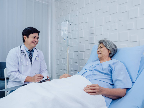 Smiling Asian doctor in white suit take notes while discussing happy Asian elderly, senior woman patient who lying on bed with receiving saline solution in hospital. Health care and medical concept. - Foto, Imagen