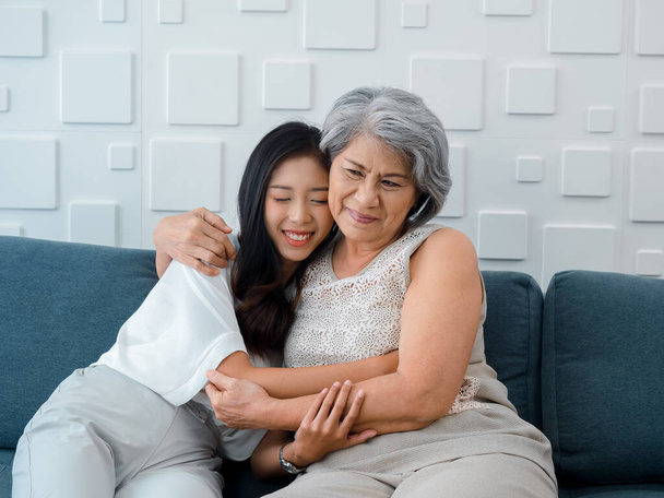 Portret szczęśliwego azjatyckiego seniora, matki lub dziadka siwe włosy obejmujące jej piękną córkę lub wnuka uśmiechające się i zamykające oczy z poczuciem miłości, podczas gdy siedzieć na szarej kanapie w salonie w domu. - Zdjęcie, obraz