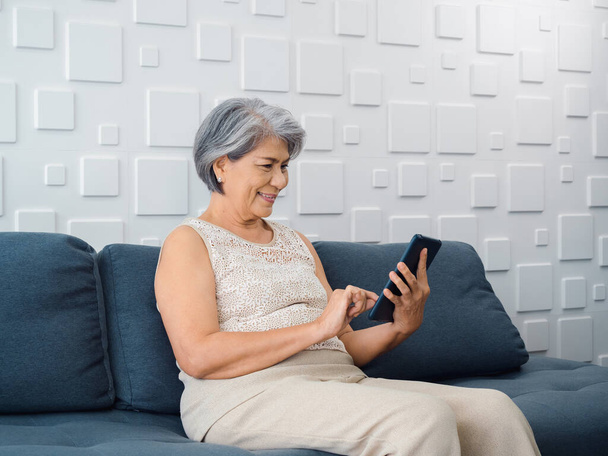 Ευτυχισμένη Ασιάτισσα ηλικιωμένη γυναίκα κάθεται στον καναπέ, κρατώντας και κοιτάζοντας την οθόνη του κινητού τηλεφώνου στο χέρι στο σαλόνι. Ηλικιωμένη γυναίκα που χρησιμοποιεί έξυπνη συσκευή, υπολογιστή στο σπίτι. Οι ηλικιωμένοι χρησιμοποιούν την τεχνολογία με ευκολία. - Φωτογραφία, εικόνα