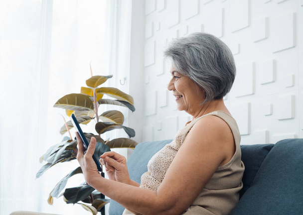 Ευτυχισμένη Ασιάτισσα ηλικιωμένη γυναίκα κάθεται στον καναπέ, κρατώντας και κοιτάζοντας ψηφιακή οθόνη tablet στο χέρι στο σαλόνι. Ηλικιωμένη γυναίκα που χρησιμοποιεί έξυπνη συσκευή, υπολογιστή στο σπίτι. Οι ηλικιωμένοι χρησιμοποιούν την τεχνολογία με ευκολία. - Φωτογραφία, εικόνα