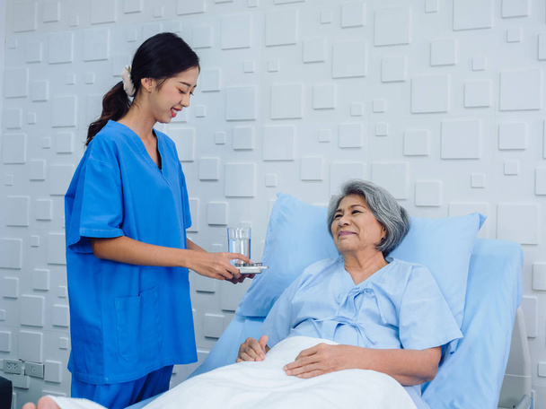 Ystävällinen hymyilevä nuori aasialainen sairaanhoitaja sinisessä pensaikossa pillereiden tarjotin iäkkäille potilaille, jotka makaavat sängyllä sairaalahuoneessa, ottaen lääkkeitä tai vitamiinilisiä, vanhempi terveydenhuolto ja lääketiede. - Valokuva, kuva