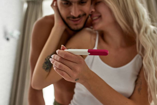 Selective focus of pregnancy test in hands of caucasian girl watching her middle eastern boyfriend. Blurred young multiethnic couple. Family creation and relationship. Maternity and parenthood - Foto, afbeelding