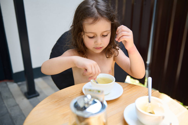 Beautiful Caucasian little girl, sitting at a table and using a tea spoon, stirring sugar in the white teacup with healthy antioxidant herbal tea - Photo, image