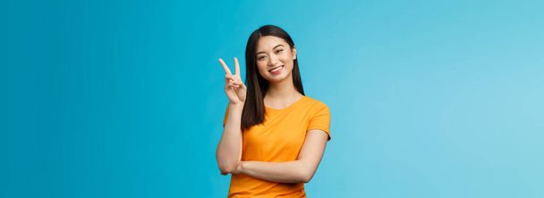 Optimistic cute tender asian young millennial girl send positive vibes, tilt head show peace, victory sign, smiling broadly, wish fantastic holidays, stand blue background joyful upbeat. - Photo, Image