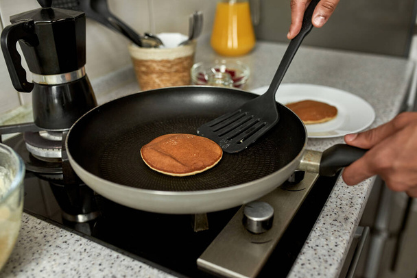 Partial view of man cooking pancake with kitchen spatula on pan on electric stove at home kitchen. Concept of domestic lifestyle. Idea of healthy eating. Modern blurred apartment. Daytime - Foto, afbeelding
