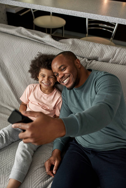 Top view of adult father and little son taking selfie on smartphone on sofa at home. Young black family lifestyle and relationship. Domestic leisure and entertainment. Fatherhood and parenting - 写真・画像