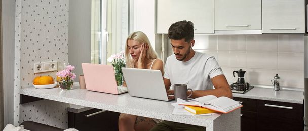 Young multiethnic couple using and working on laptops at table at home kitchen. Middle eastern man and blonde caucasian girl. Remote work and freelance. Domestic lifestyle. Modern spacious flat - Zdjęcie, obraz
