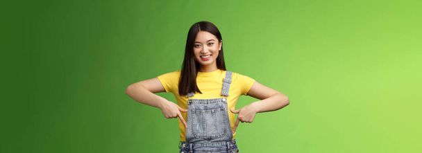 friendly outgoing cute asian young woman show fingers down, introduce new promo app, smiling joyfully sharing link, recommend product using brand, grinning toothy, green background. Copy space - Фото, изображение