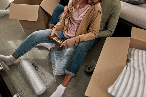Cropped top view of smiling young couple resting and looking family photo on floor during packing things in cardboard box. Man and woman moving to new apartment. Concept of home relocation - Fotoğraf, Görsel