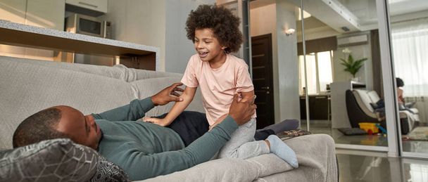 Smiling little boy sitting near lying adult father on sofa at home. Black man and child spending time together and looking at each other. Family relationship. Domestic lifestyle. Modern flat interior - Foto, Bild