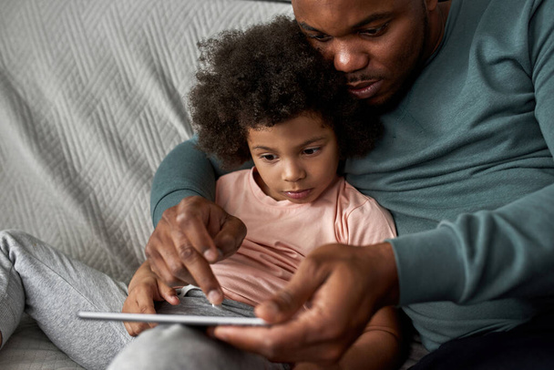 Partial adult father and little son using and watching digital tablet on sofa at home. Young black family lifestyle and relationship. Domestic leisure and entertainment. Fatherhood and parenting - Foto, Bild