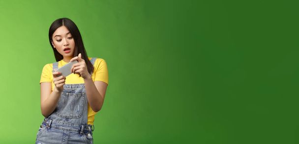 Huoleton nörtti söpö aasialainen brunette tuhlata aikaa yliopiston kampuksella, pelaa online älypuhelin peli, näyttää huvittunut viihdyttävä puhelinnäyttö, kallistaa kehon liikkua merkki, avoin suu innoissaan. - Valokuva, kuva