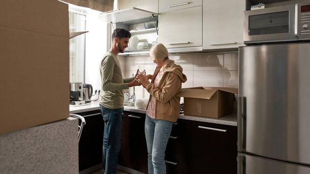 Smiling middle eastern man giving two glasses to his blonde wife or girlfriend for packing in cardboard box. Concept of home moving. Young couple. Interior of modern spacious apartment - Fotó, kép