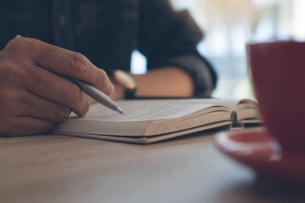 Homem segurando caneta e livro de leitura com xícara de café na mesa de madeira - Foto, Imagem