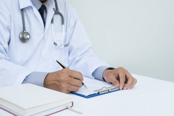 Detailní záběr mužského lékaře vyplňující pacientův dokument na schránce černým perem na bílém stole v nemocnici. - Fotografie, Obrázek