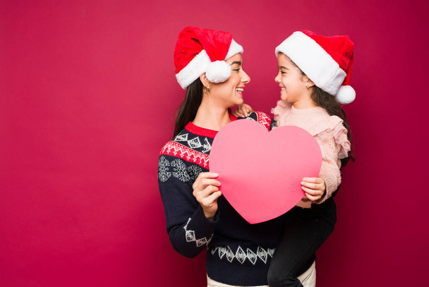 Ελκυστική στοργική μαμά που κουβαλάει την κόρη της γελώντας φορώντας χριστουγεννιάτικα πουλόβερ και καπέλα κρατώντας μια καρδιά  - Φωτογραφία, εικόνα