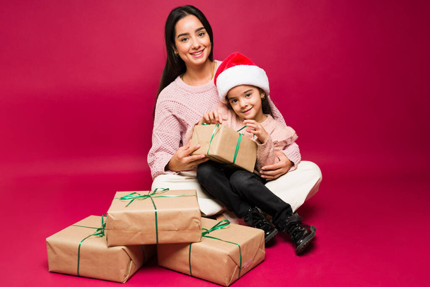 Lächelnde Mutter und kleine Tochter blicken beim gemeinsamen Öffnen der Weihnachtsgeschenke in die Kamera - Foto, Bild