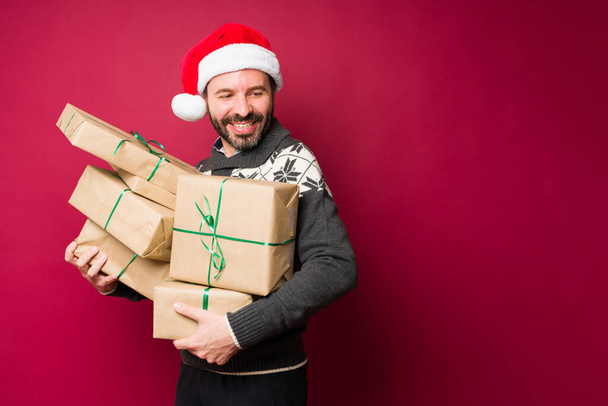 Attractive latin man with a santa hat laughing and enjoying giving a receiving a lot of christmas presents for family and friends - Photo, Image