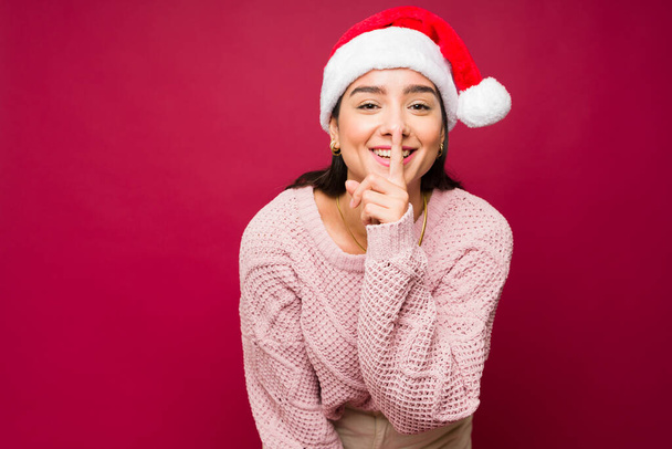 Adorável bela jovem usando um chapéu de Papai Noel colocando um dedo em seus lábios e pedindo para manter um segredo para as férias - Foto, Imagem