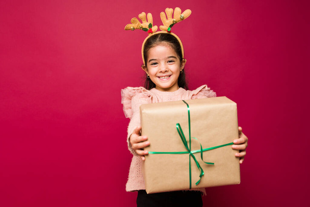 Adorable hispanic girl making eye contact while smiling and giving friends or family a Christmas gift - Foto, afbeelding