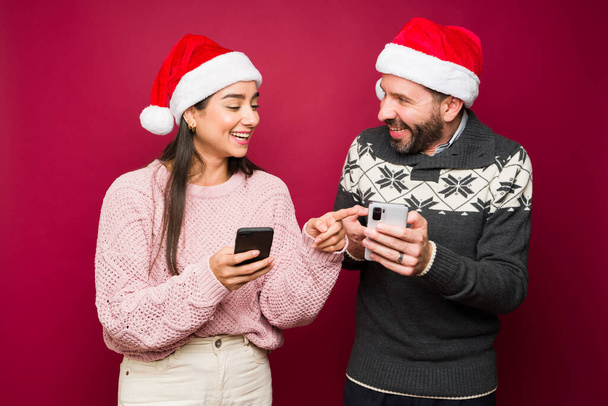 Couple joyeux riant tout en envoyant des SMS à la famille et aux amis pour les vacances de Noël et en utilisant les médias sociaux  - Photo, image