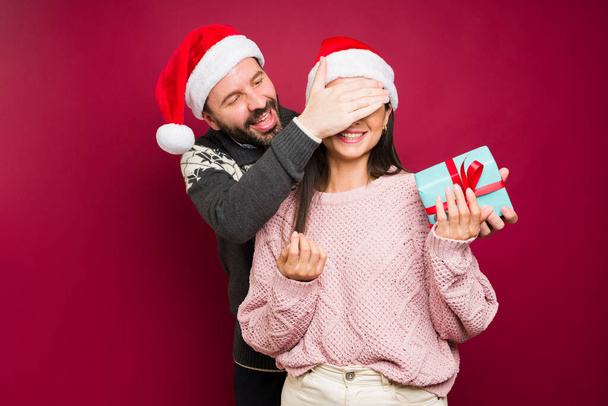 Χαρούμενος άντρας με καπέλο Σάντα που καλύπτει τα μάτια μιας ενθουσιασμένης νεαρής γυναίκας για να της κάνει έκπληξη με χριστουγεννιάτικο δώρο. - Φωτογραφία, εικόνα