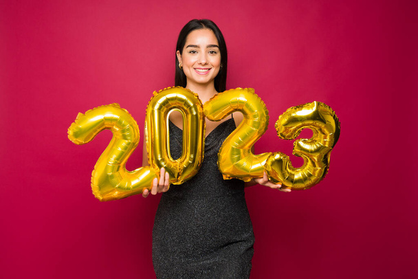 Gorgeous young woman at a party wearing a black dress showing 2023 balloons to celebrate new year's eve - Foto, afbeelding