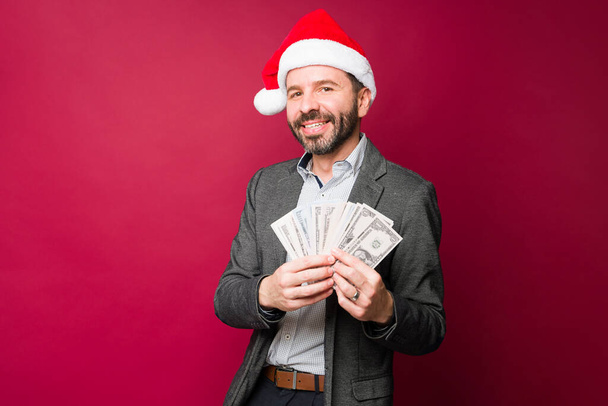 Riche homme hispanique souriant et regardant heureux tout en ayant beaucoup d'argent pendant les vacances de Noël - Photo, image